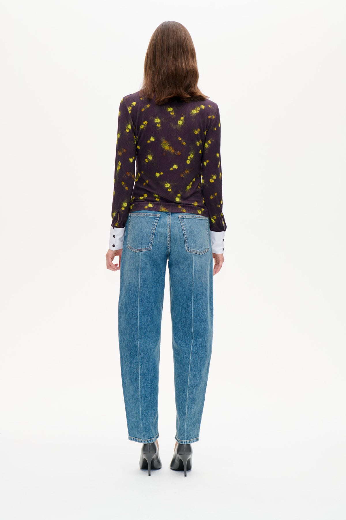 Rückenansicht eines Models, das ein langärmeliges dunkelviolettes Oberteil mit vereinzeltem gelb-grünem Blumenmuster trägt, dazu weiße Manschetten und eine blaue Jeans.