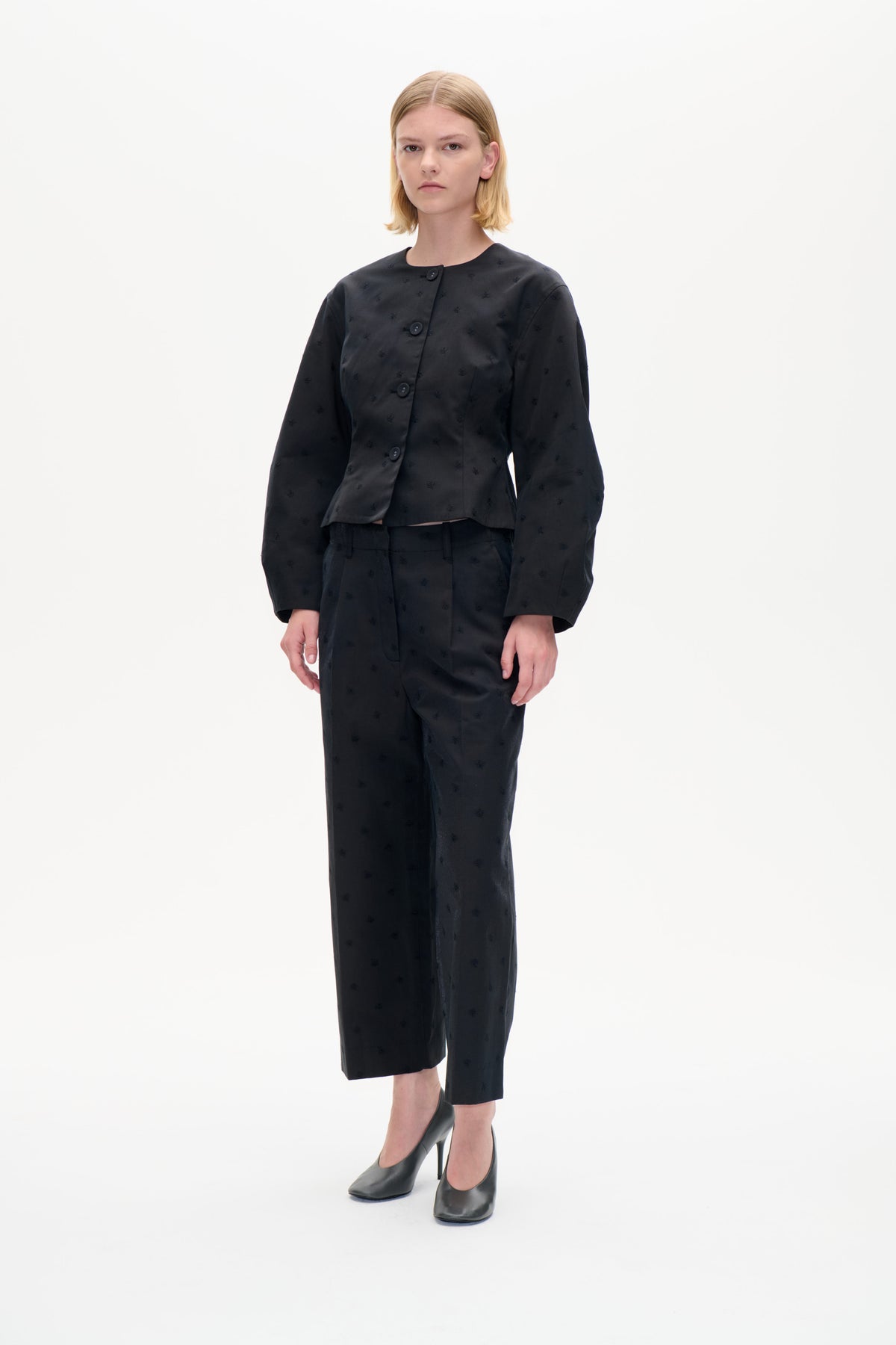 Model trägt eine anthrazit-schwarze Jacke mit kleinen Stickmotiven und asymmetrischer Knopfleiste, kombiniert mit passenden, kurz geschnittenen Hosen.
