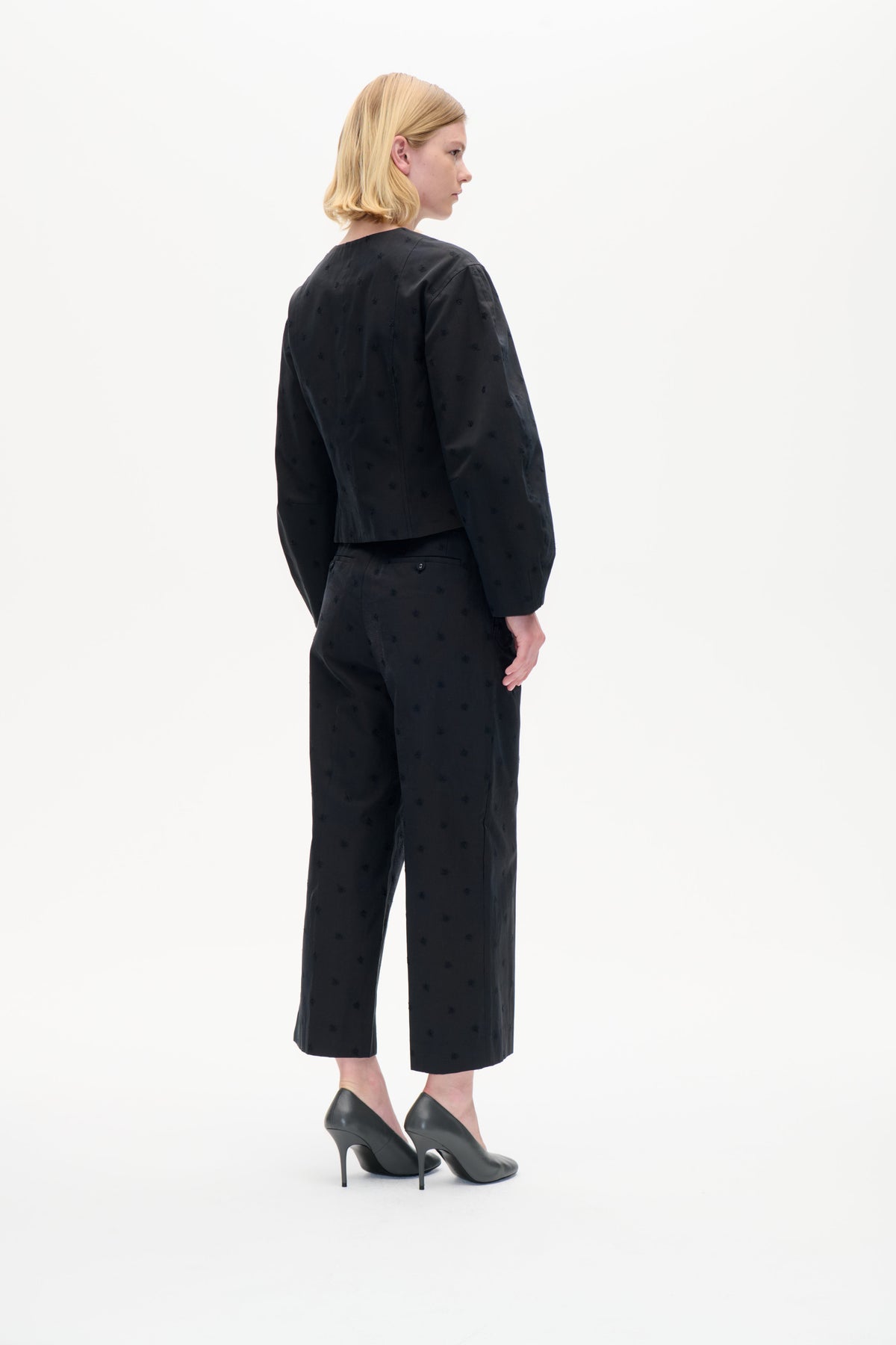 Model von hinten im Profil trägt ein schwarzes, langärmliges Top und eine passende, verkürzte Hose mit dezenten Ton-in-Ton-Motiven.