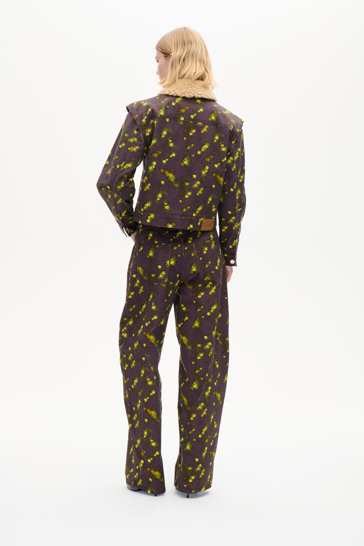 Rückenansicht eines Models, das eine dunkelpflaumenfarbene Jacke mit leuchtend gelb-grünem Blumenmuster und beigem, strukturiertem Kragen trägt, kombiniert mit passender Hose.
