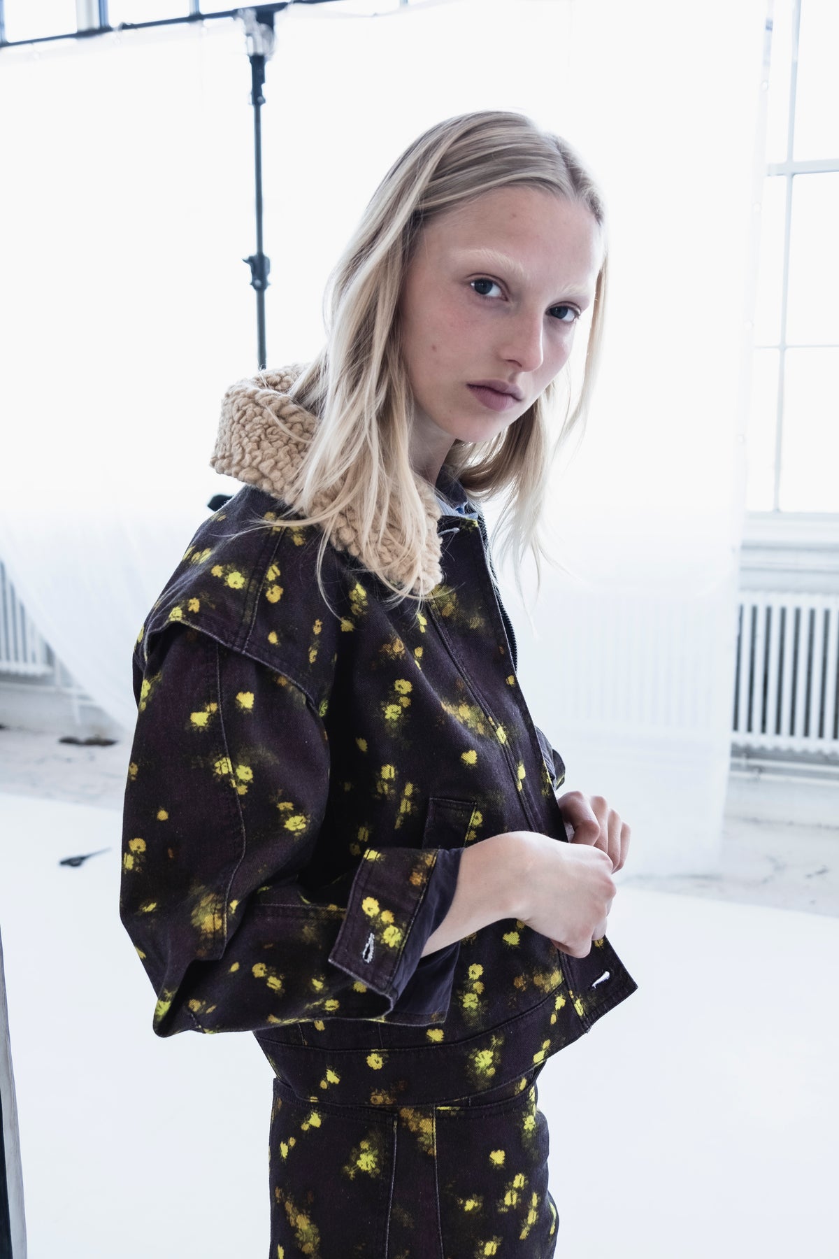 Kampagnen-Nahaufnahme eines Models in einer dunklen Jacke mit verstreuten gelben Blumenmotiven und einem dicken beigefarbenen Strickschal.