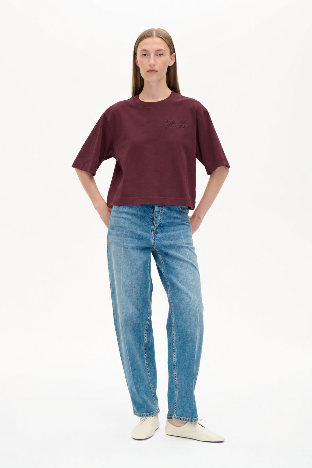 Vorderansicht eines Models, das ein burgunderfarbenes T‑Shirt mit kurzen Ärmeln und tonalem Brustlogo sowie hellblaue, gemusterte Wide‑Leg-Jeans trägt.