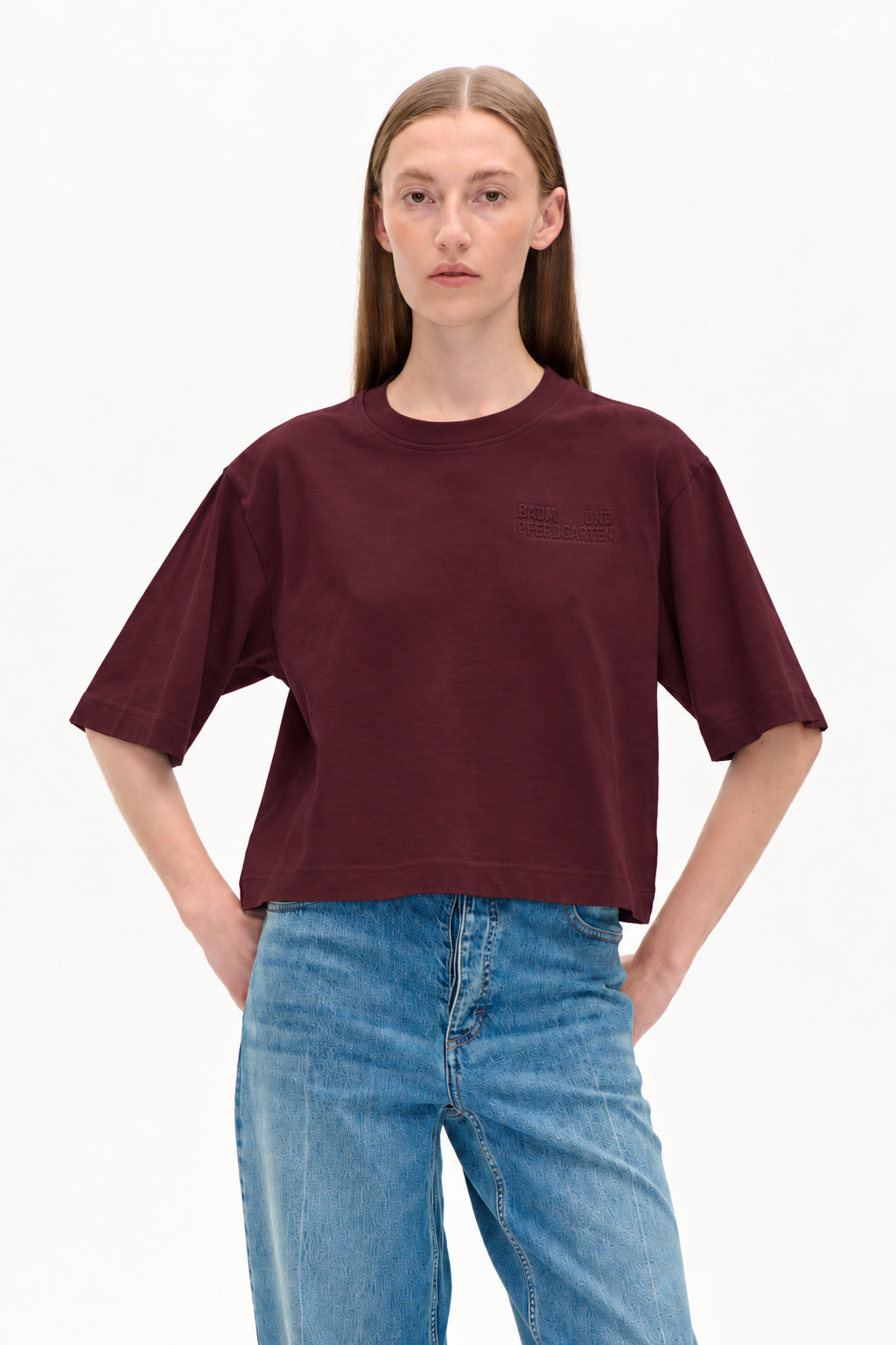 Model trägt ein kurz geschnittenes bordeauxfarbenes T-Shirt mit kurzen Ärmeln und geprägtem Brustlogo sowie eine hellblaue, gemusterte Jeans.