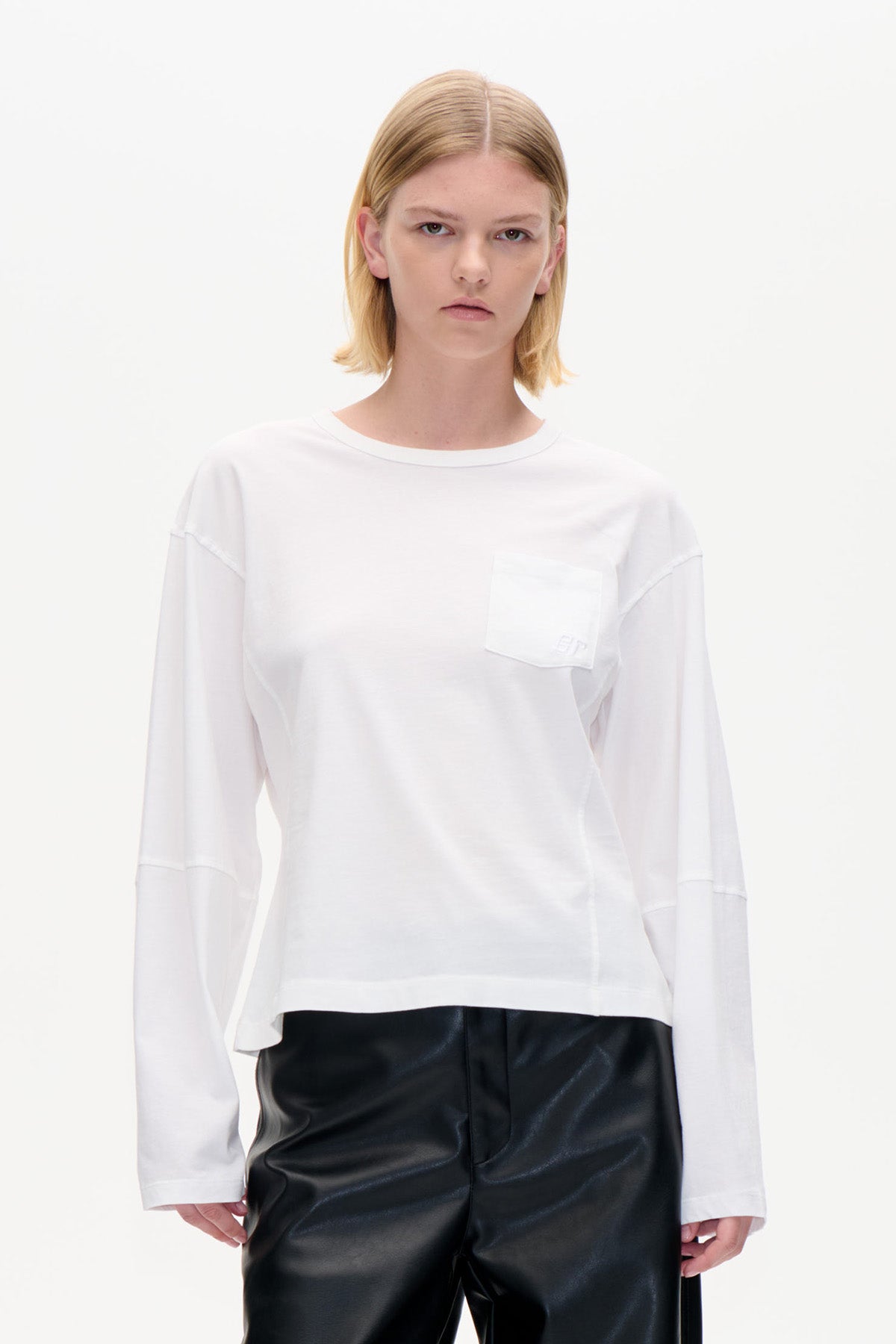 Vorderansicht eines weißen, langärmeligen Tops mit Rundhalsausschnitt und Brusttasche, gestylt mit einer schwarzen Hose.