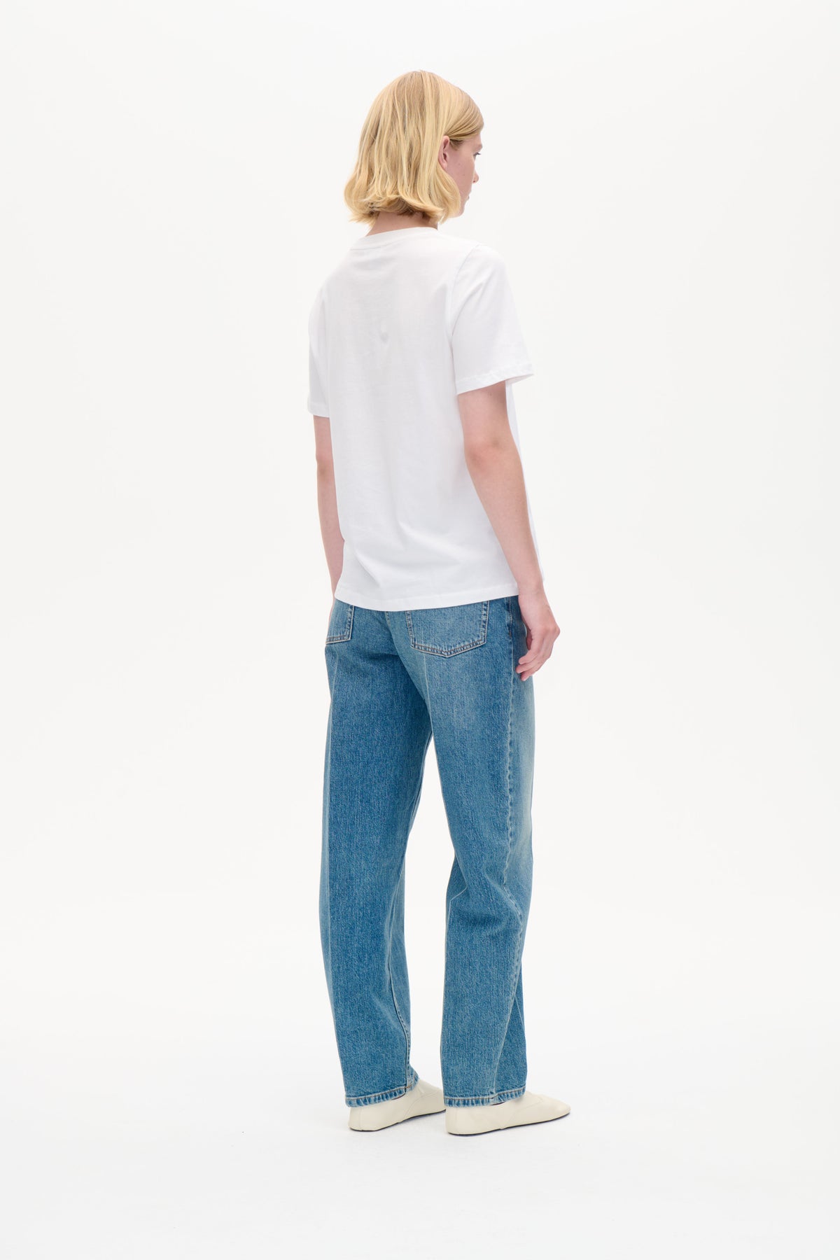 Rückansicht eines Models, das ein schlichtes weißes kurzärmliges T-Shirt, blaue Jeans und weiße Schuhe vor weißem Hintergrund trägt.