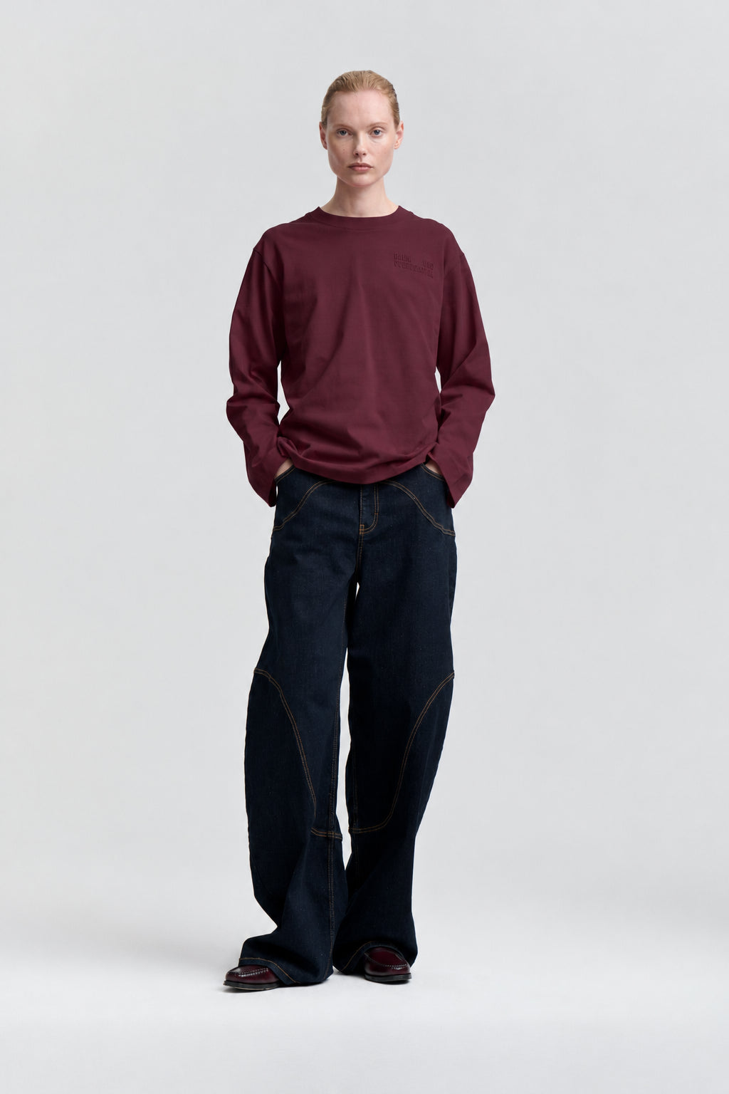 Frontalaufnahme eines Models in einem burgunderfarbenen, langärmligen Sweatshirt und weiten, dunkelindigo Jeans mit Kontrastnähten.