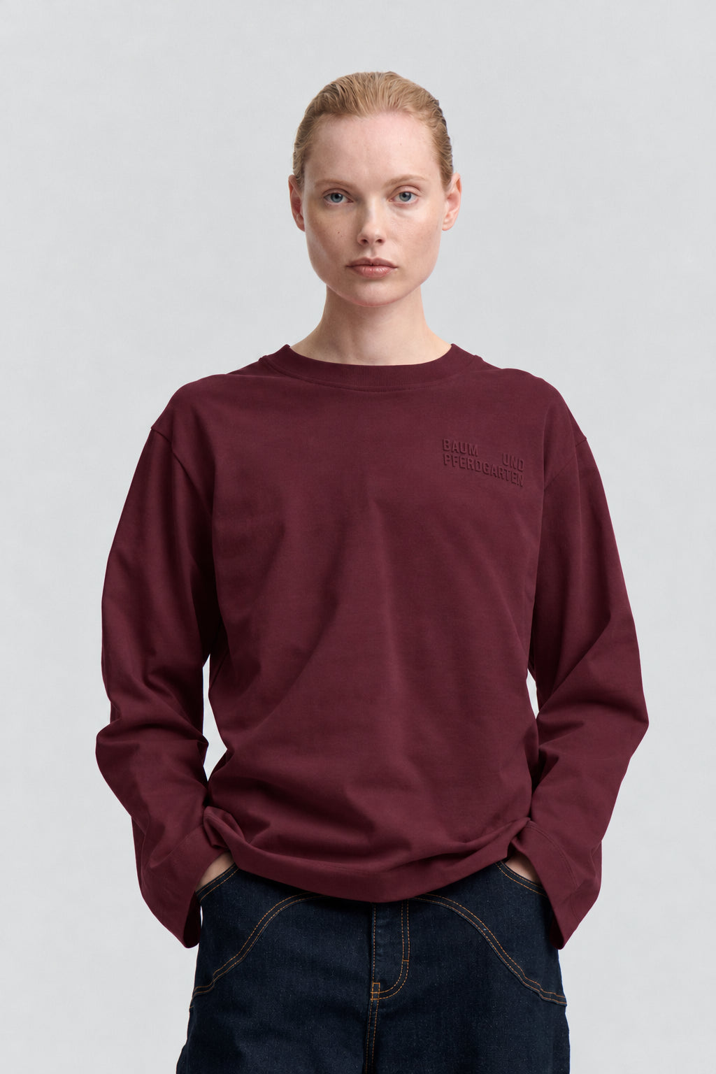 Frontalansicht eines burgunderroten Sweatshirts mit Rundhalsausschnitt, langen Ärmeln und einem dezenten tonalen Schriftzug auf der Brust, gestylt mit dunklen Jeans.