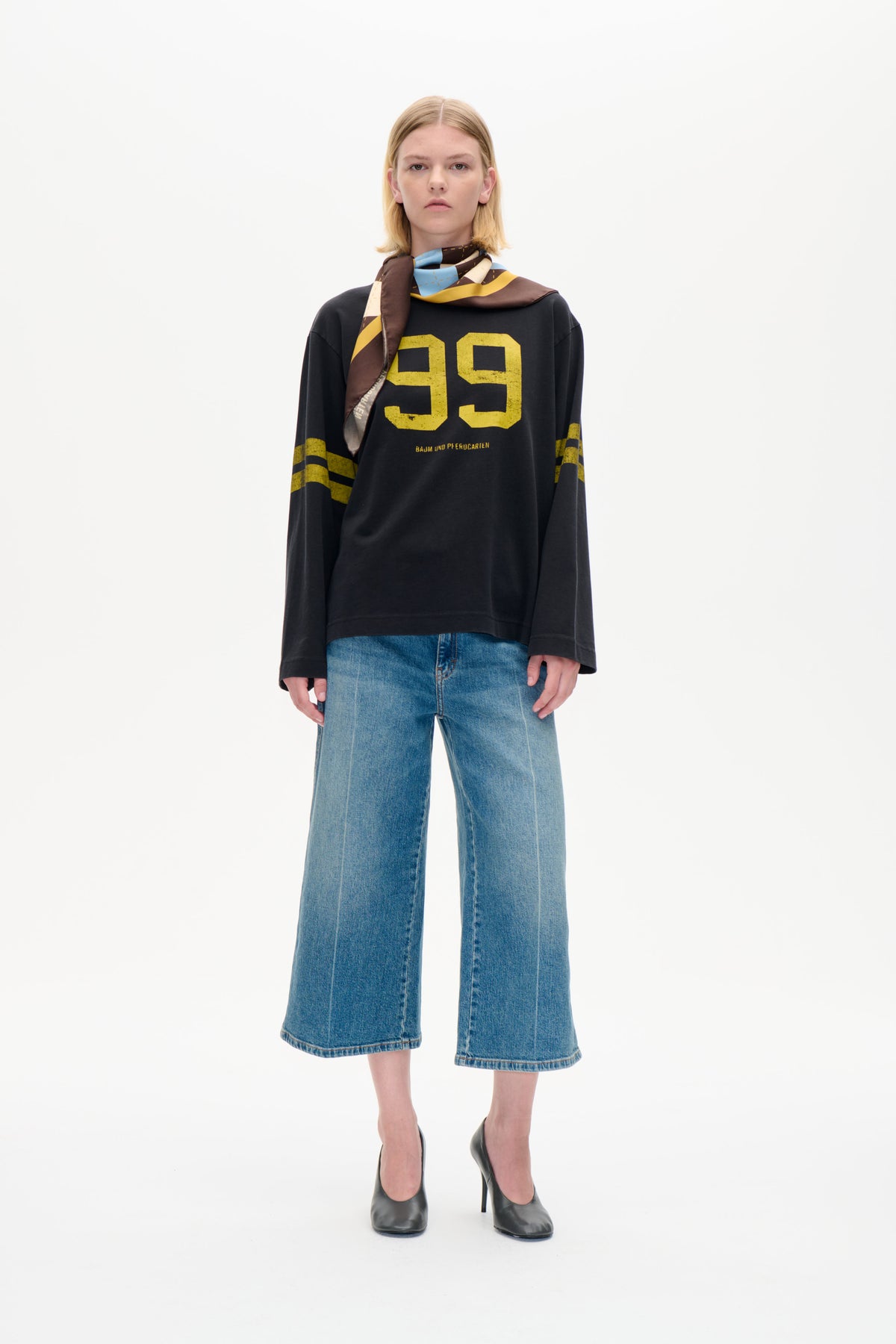 Model trägt ein anthrazitfarbenes, langärmeliges Sweatshirt mit großem gelben 99-Aufdruck, eine weit geschnittene blaue Jeans und einen mehrfarbigen Schal mit geometrischem Muster.