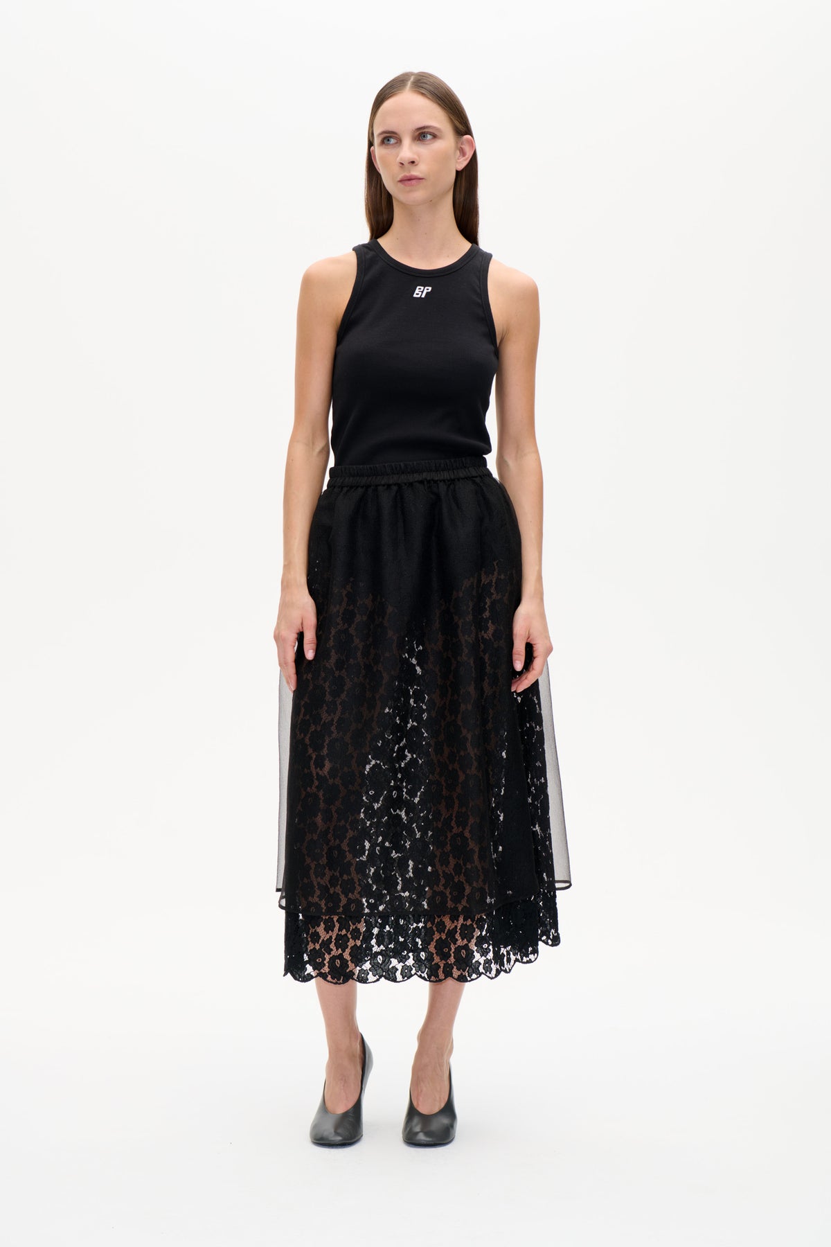 Vorderansicht eines Models, das ein schwarzes, geripptes ärmelloses Top und einen schwarzen, floralen Spitzenrock mit transparentem Überzug und Muschelsaum trägt.