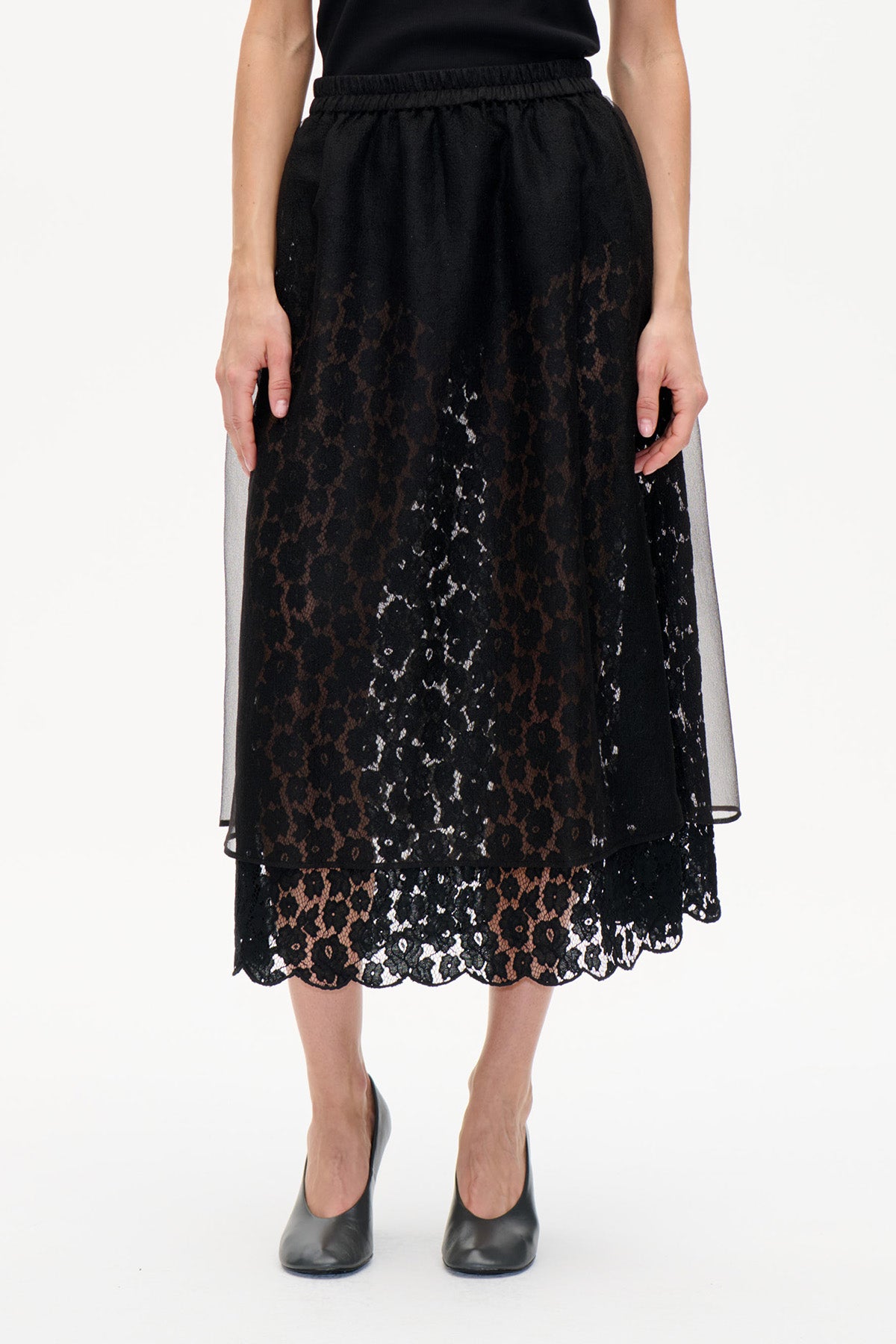 Vorderansicht eines schwarzen midi-Rocks mit floraler Spitze, geschwungenem Saum und transparentem Overlay, kombiniert mit einem schwarzen Top.