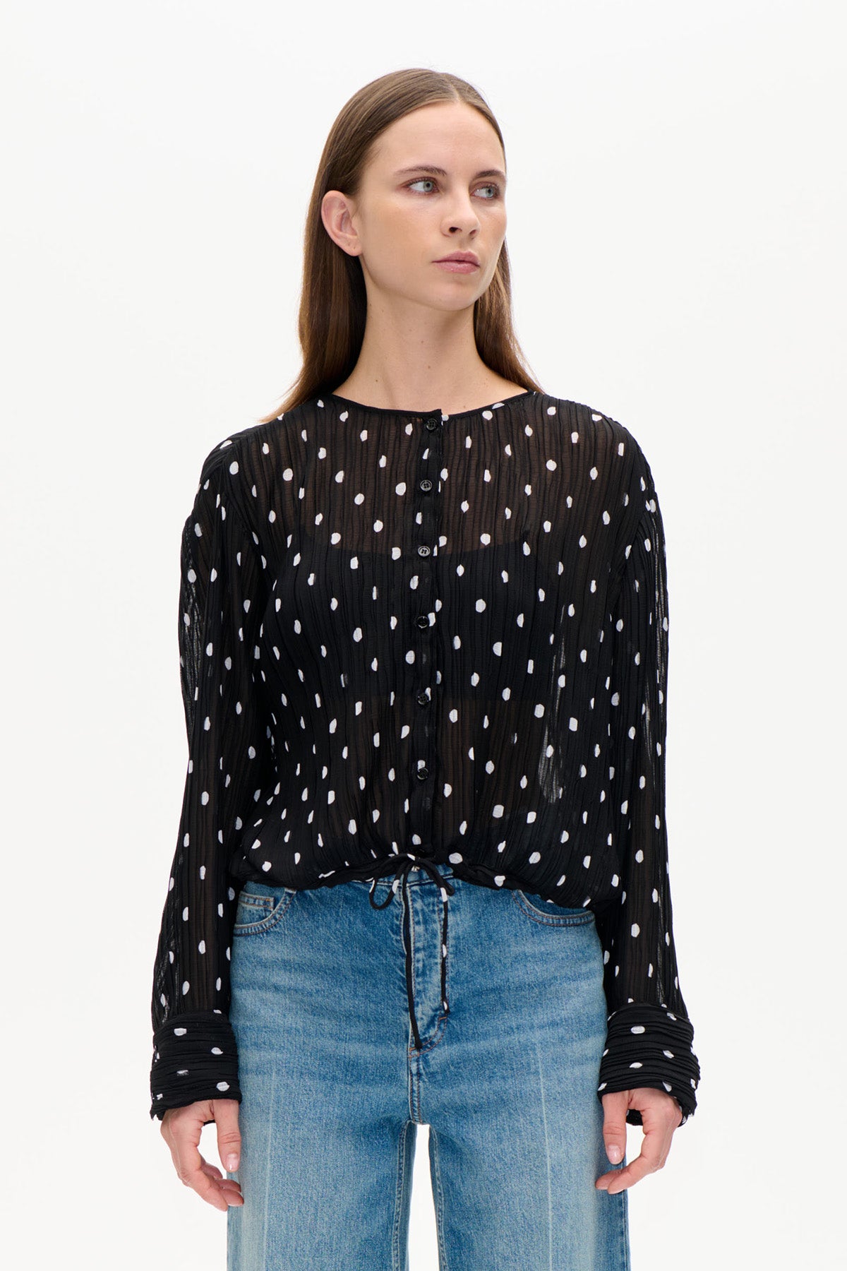 Vorderansicht einer schwarzen, transparenten, gekräuselten Bluse mit unregelmäßigen weißen Punkten, durchgehender Knopfleiste und langen Ärmeln, kombiniert mit blauen Jeans.