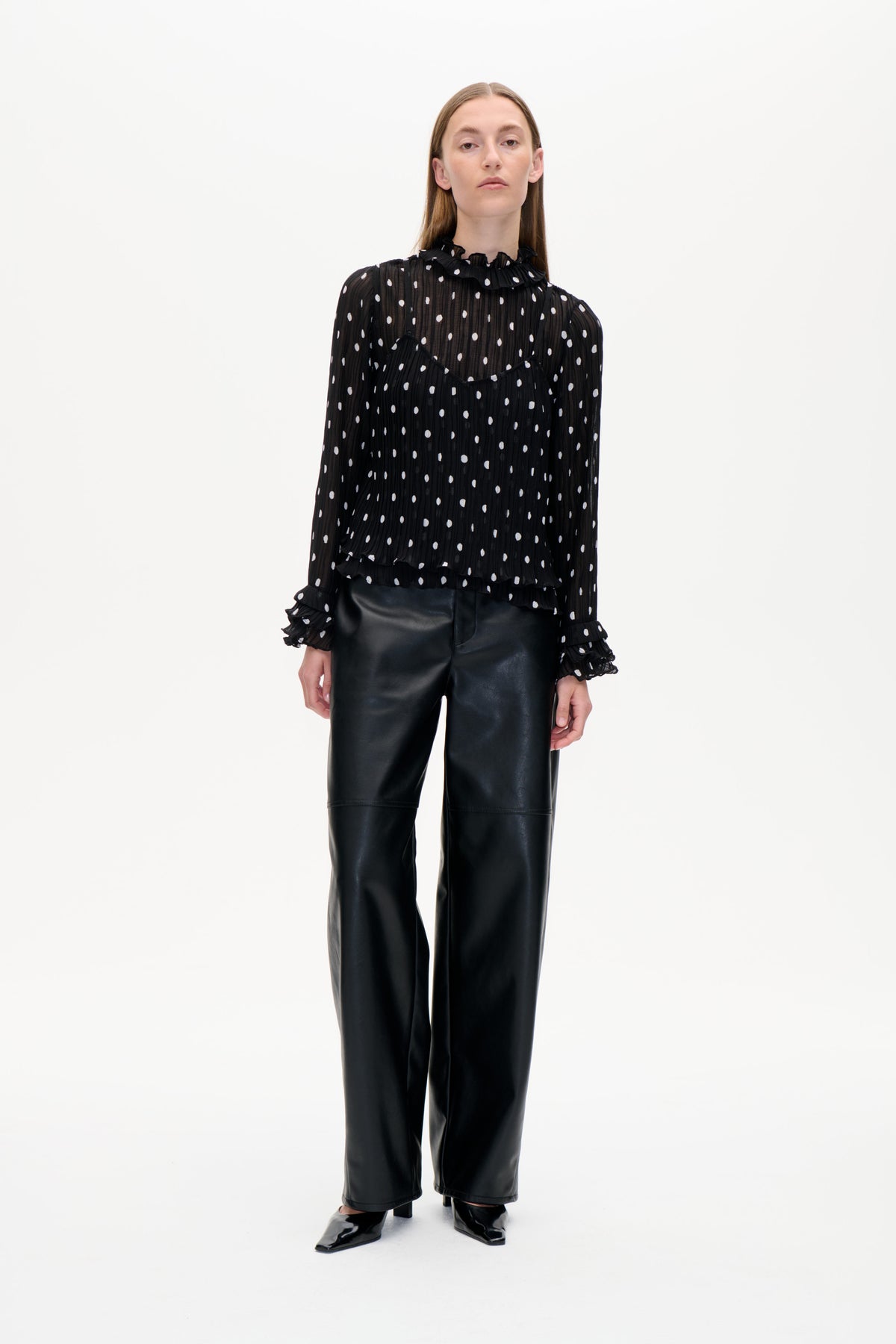 Frontal aufgenommenes Model in transparenter schwarzer Bluse mit weißen Punkten und Rüschenbesatz, kombiniert mit schwarzen, weitgeschnittenen Hosen.