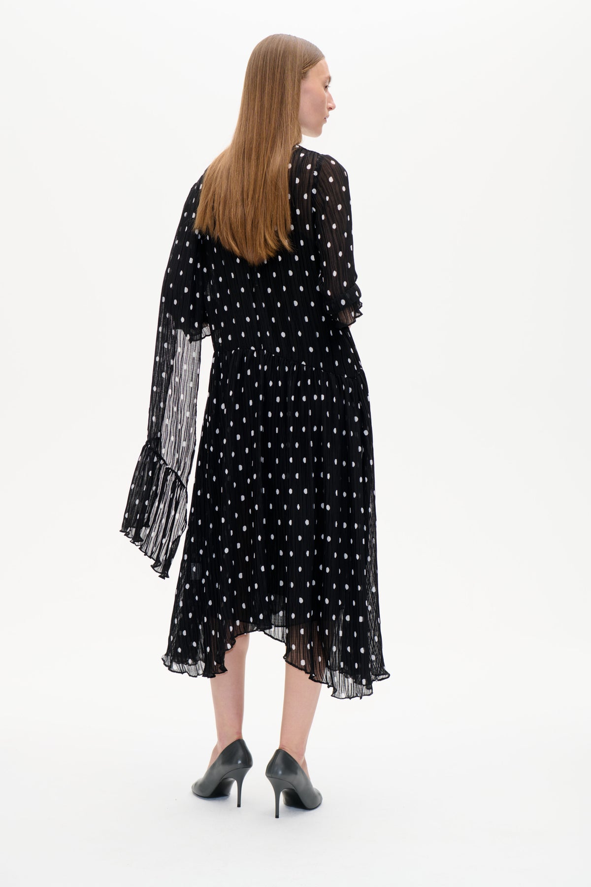 Model trägt ein schwarzes, transparentes, plissiertes Midikleid mit weißen Punkten und Rüschenärmeln, von hinten gezeigt, in schwarzen High Heels.