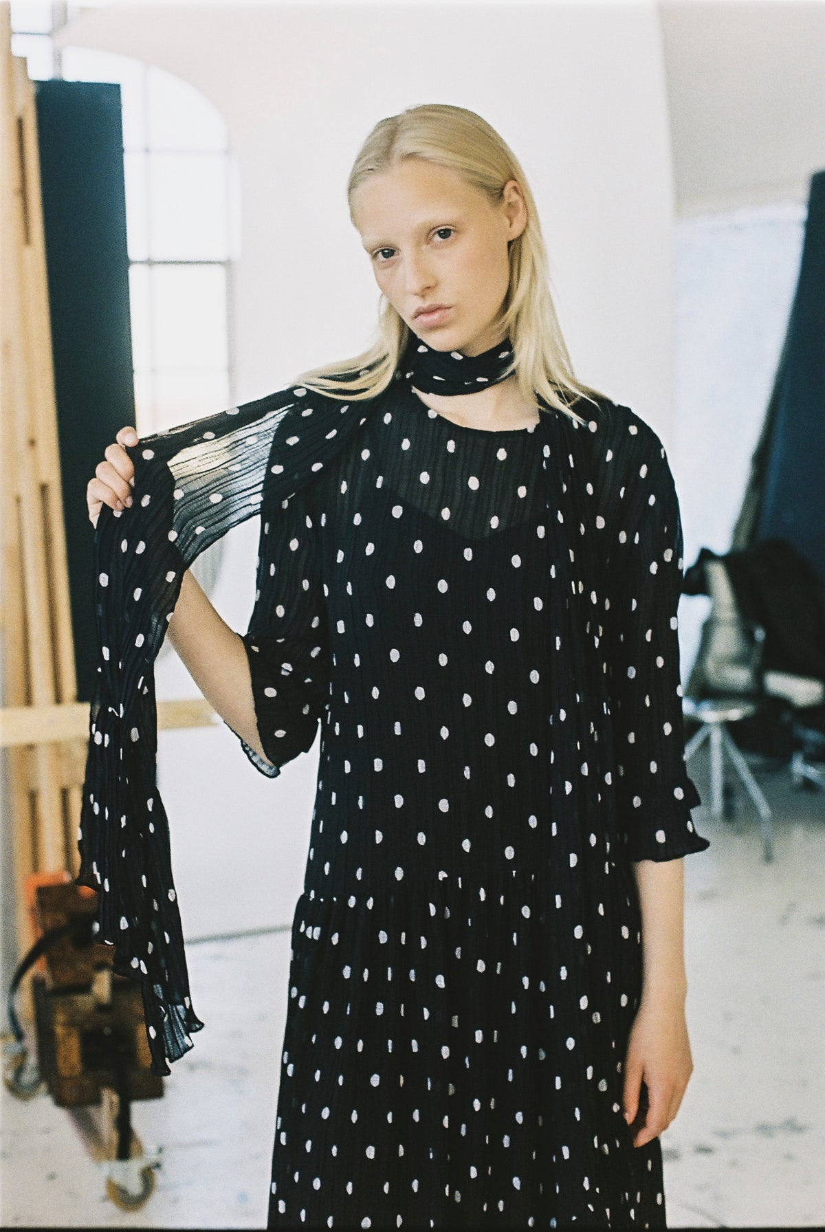 Model trägt ein schwarzes Kleid mit unregelmäßigen weißen Punkten, einem hohen Ausschnitt und transparenten, plissierten Ärmeln und hält einen Ärmel ausgestreckt.