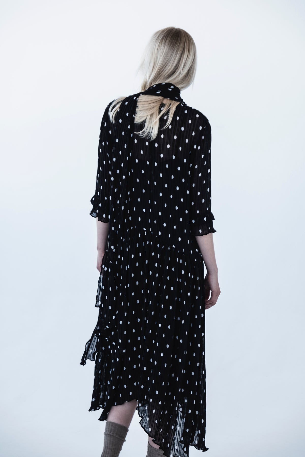 Rückenansicht eines Models, das ein schwarzes, plissiertes Kleid mit unregelmäßigen weißen Punkten, Flatterärmeln und mehrlagigem, transparentem Saum vor hellem Hintergrund trägt.