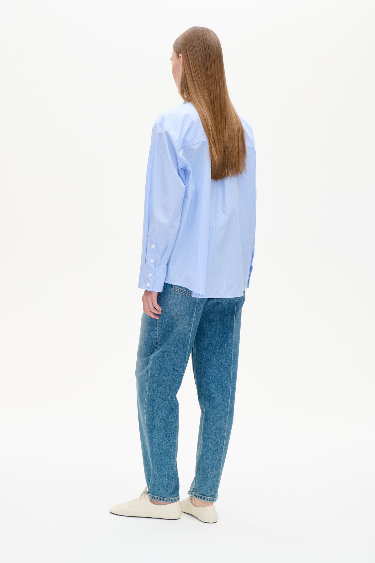 Rückenansicht eines Models in einem hellblauen, fein gestreiften Knopfhemd mit langen Ärmeln und blauen Jeans.