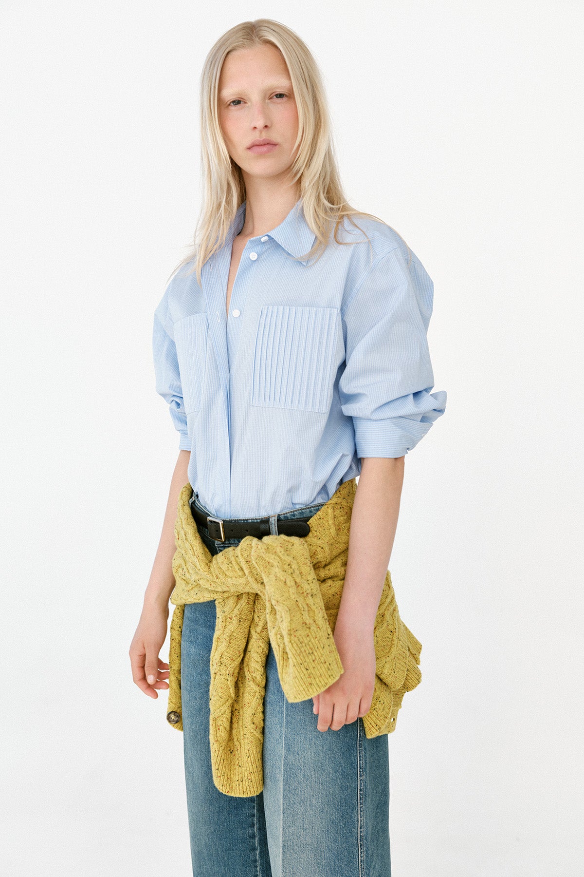 Model trägt eine hellblaue, oversized Bluse mit plissierter Brusttasche, weite blaue Jeans und einen senffarbenen Strick, in der Taille geknotet.