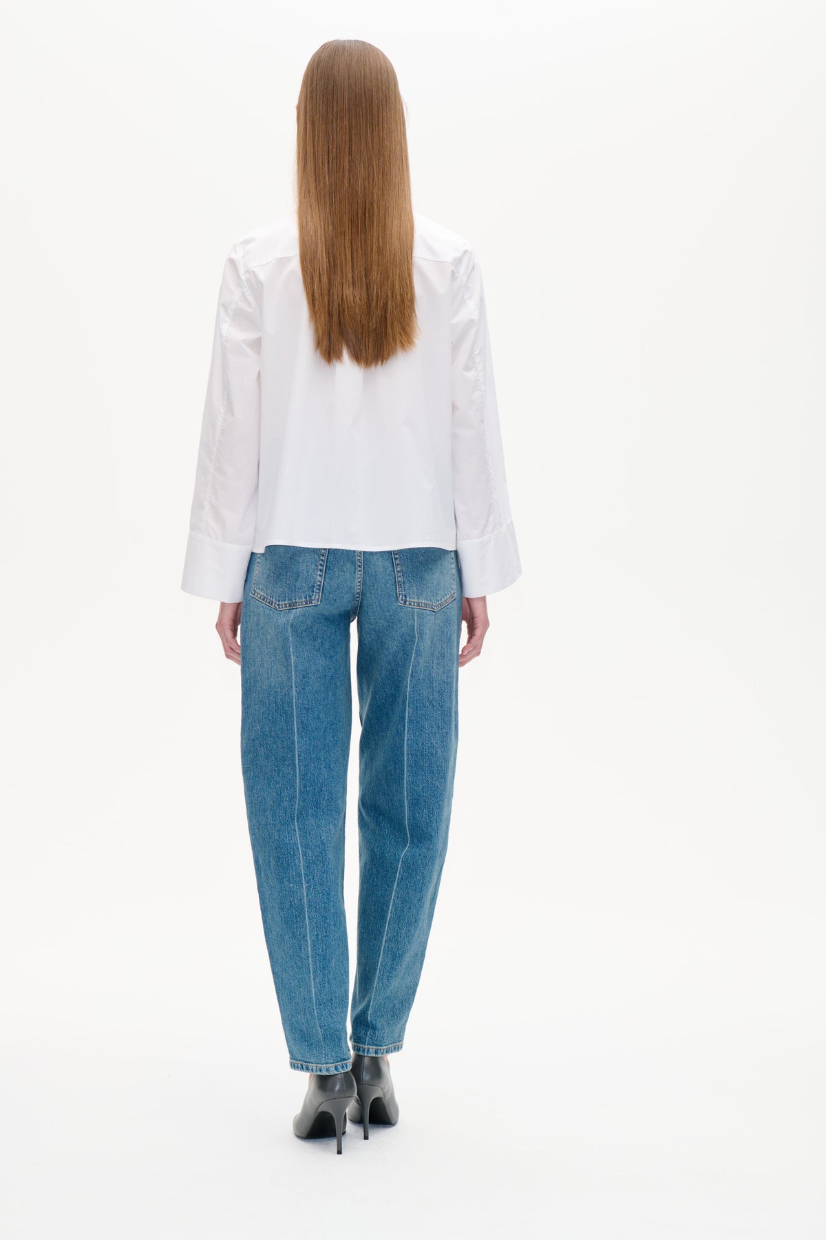 Rückansicht eines Models, das eine weiße Langarmbluse mit weiten Manschetten und blaue Jeans mit aufgesetzten Gesäßtaschen trägt.