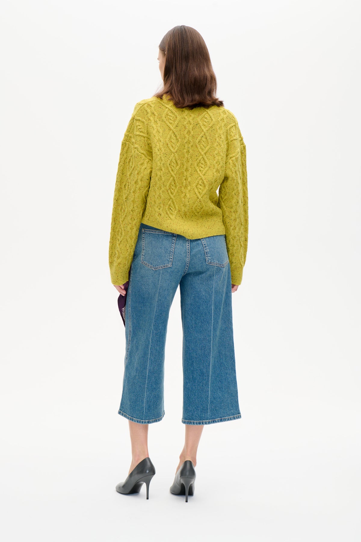 Rückenansicht eines Models, das einen gelb-grün gesprenkelten Zopfstrickpullover und eine blaue, verkürzte Jeans mit weitem Bein trägt.