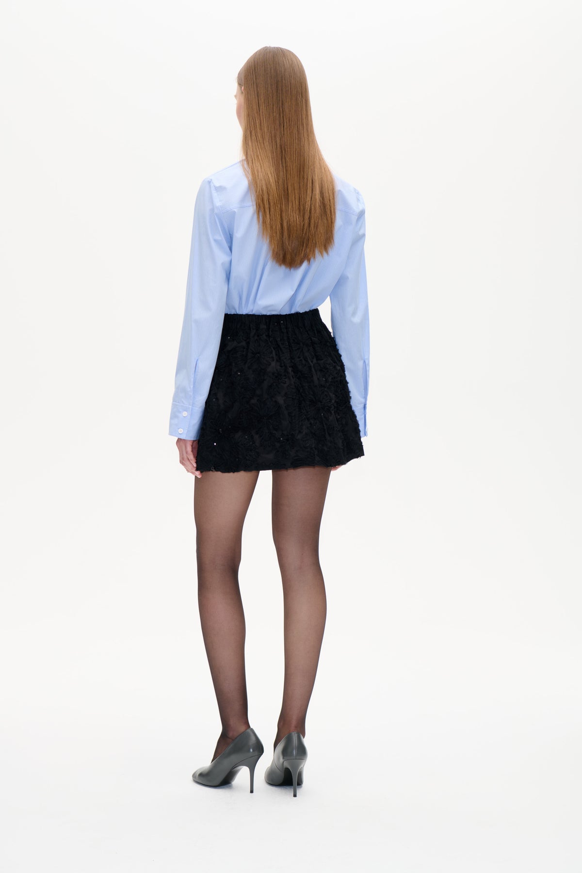 Rückenansicht eines Models, das eine hellblaue Nadelstreifenbluse und einen schwarzen, strukturierten Minirock mit Blumenmuster trägt.