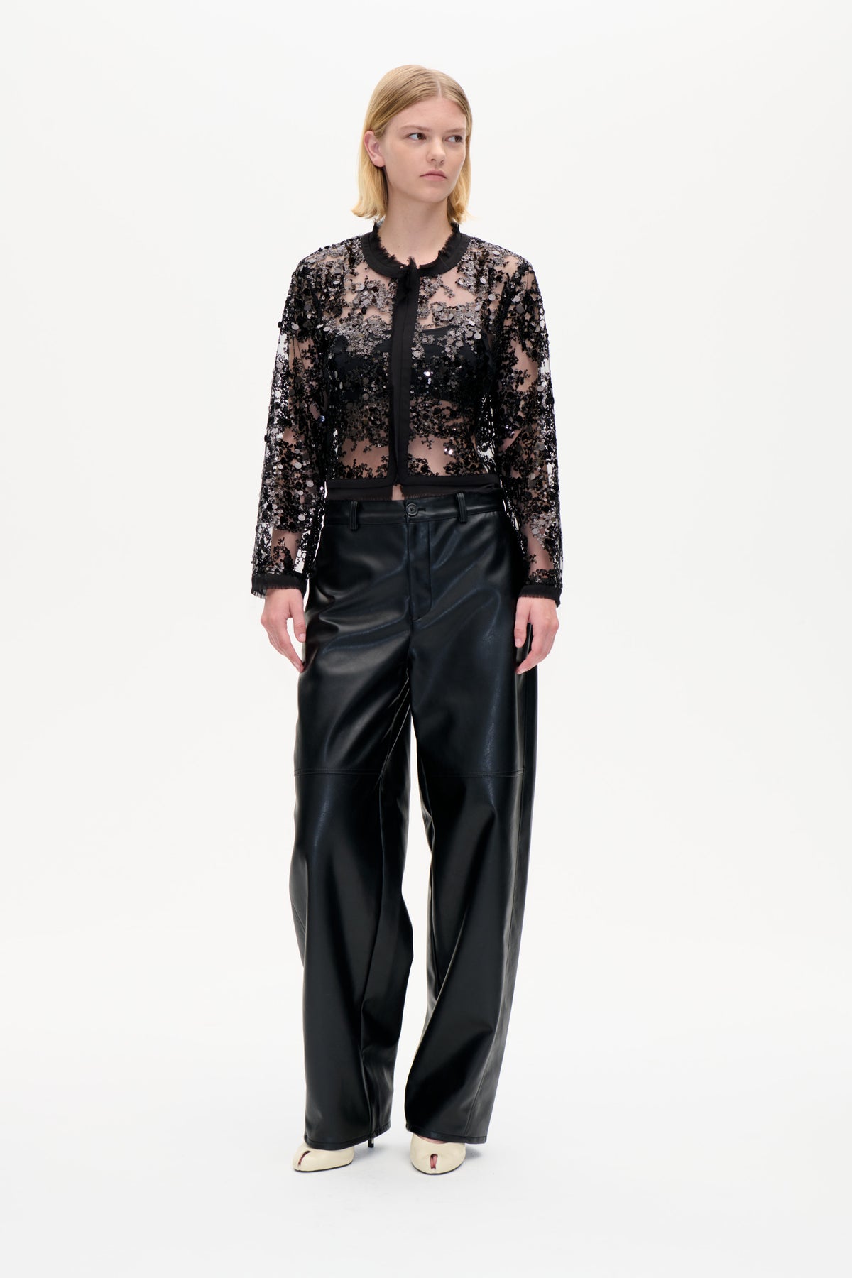 Model trägt eine transparente schwarze Paillettenbluse mit gebundenem Zierkragen und eine weite schwarze Hose, frontal vor weißem Studiohintergrund.