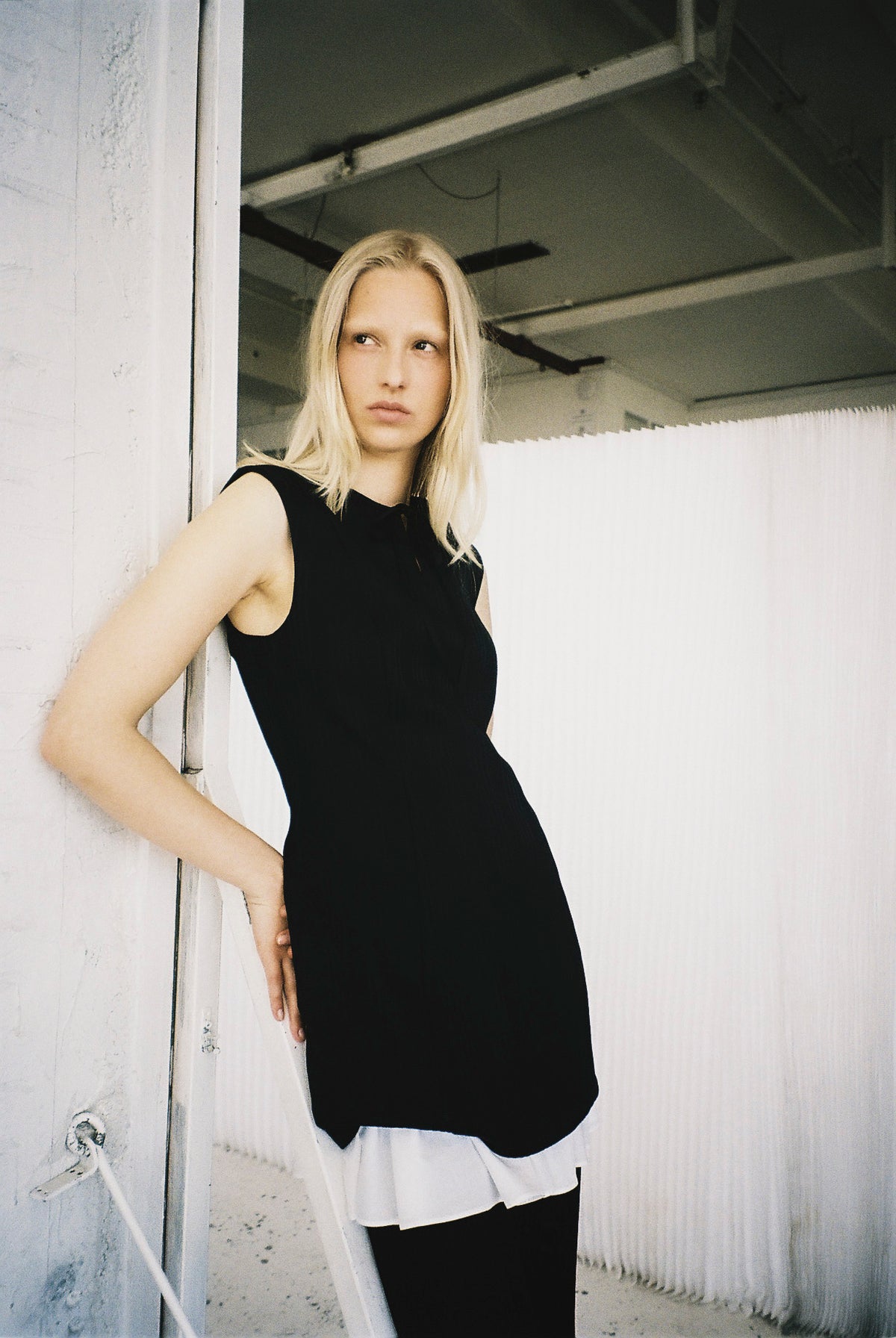 Model in einer schwarzen, ärmellosen, figurbetonten Tunika über einer weißen Bluse mit Rüschensaum, neben einer weißen Wand.