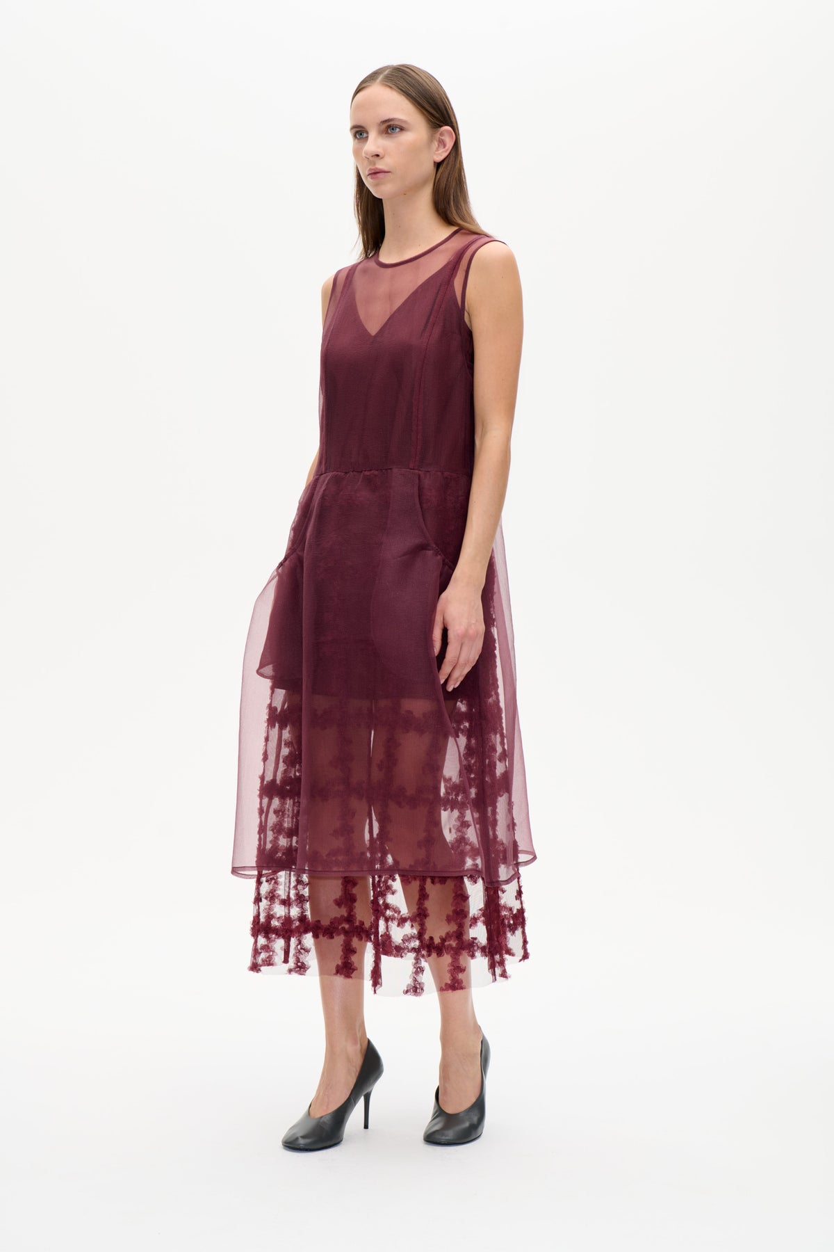 Model trägt ein pflaumenfarbenes, ärmelloses Kleid mit transparentem Überwurf und dunkler Blumenstickerei, frontal vor weißem Hintergrund.