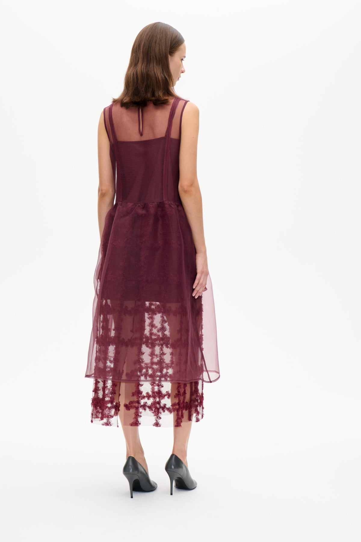 Rückansicht eines Models in einem burgunderfarbenen, ärmellosen, mehrlagigen Kleid mit transparentem Rückenteil und transparentem Rock mit floralem Muster