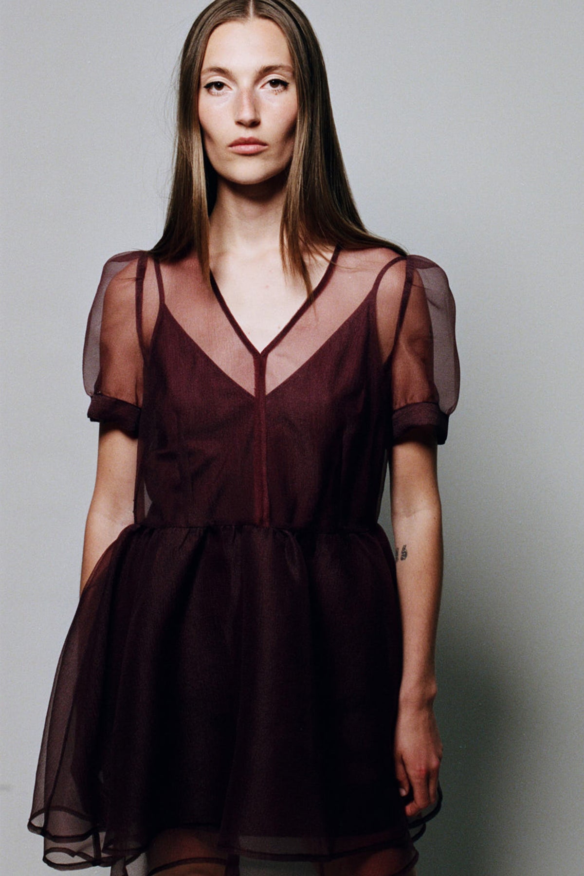 Frontalaufnahme für die Kampagne: Model trägt ein dunkles bordeauxfarbenes Kleid mit V‑Ausschnitt, kurzen transparenten Ärmeln und einem fließenden, transparenten Überwurf.