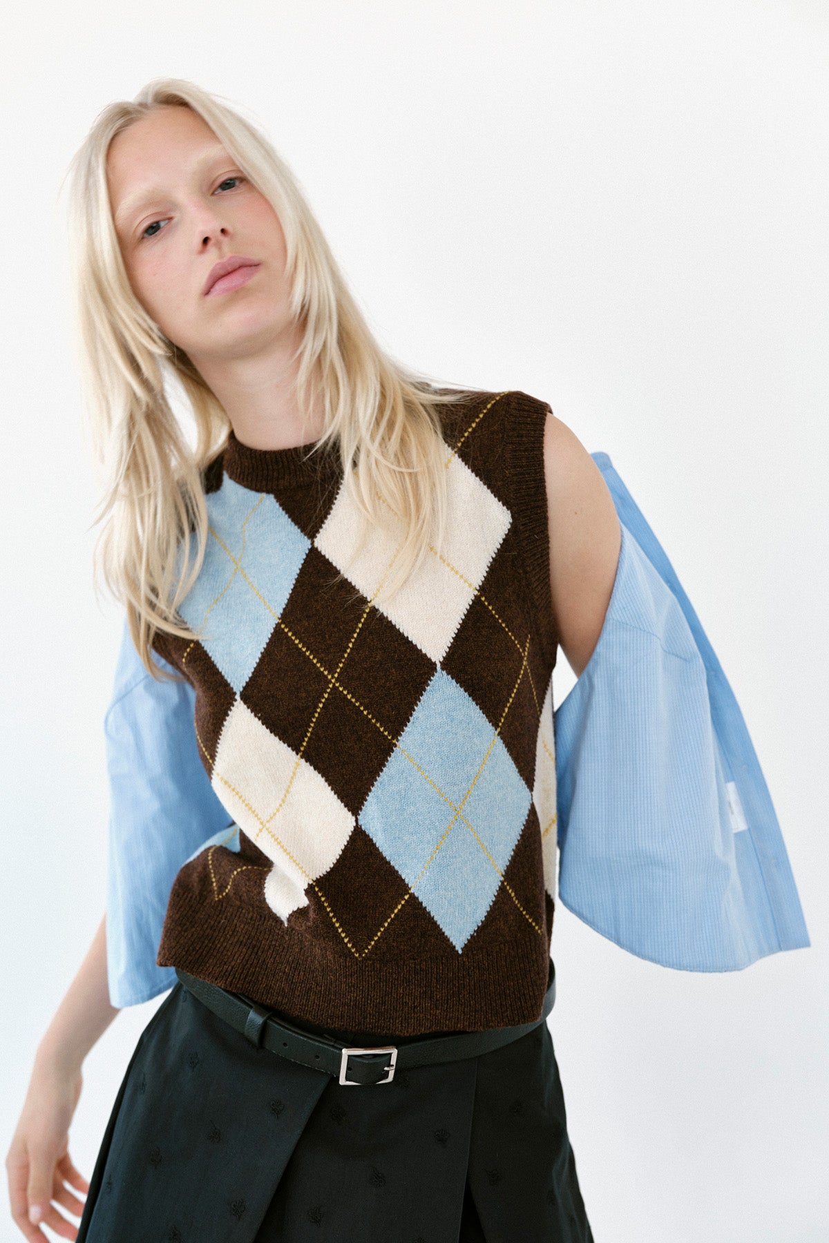 Model trägt eine braune ärmellose Strickweste mit blauen und cremefarbenen Argyle-Rauten über einer hellblauen Bluse.