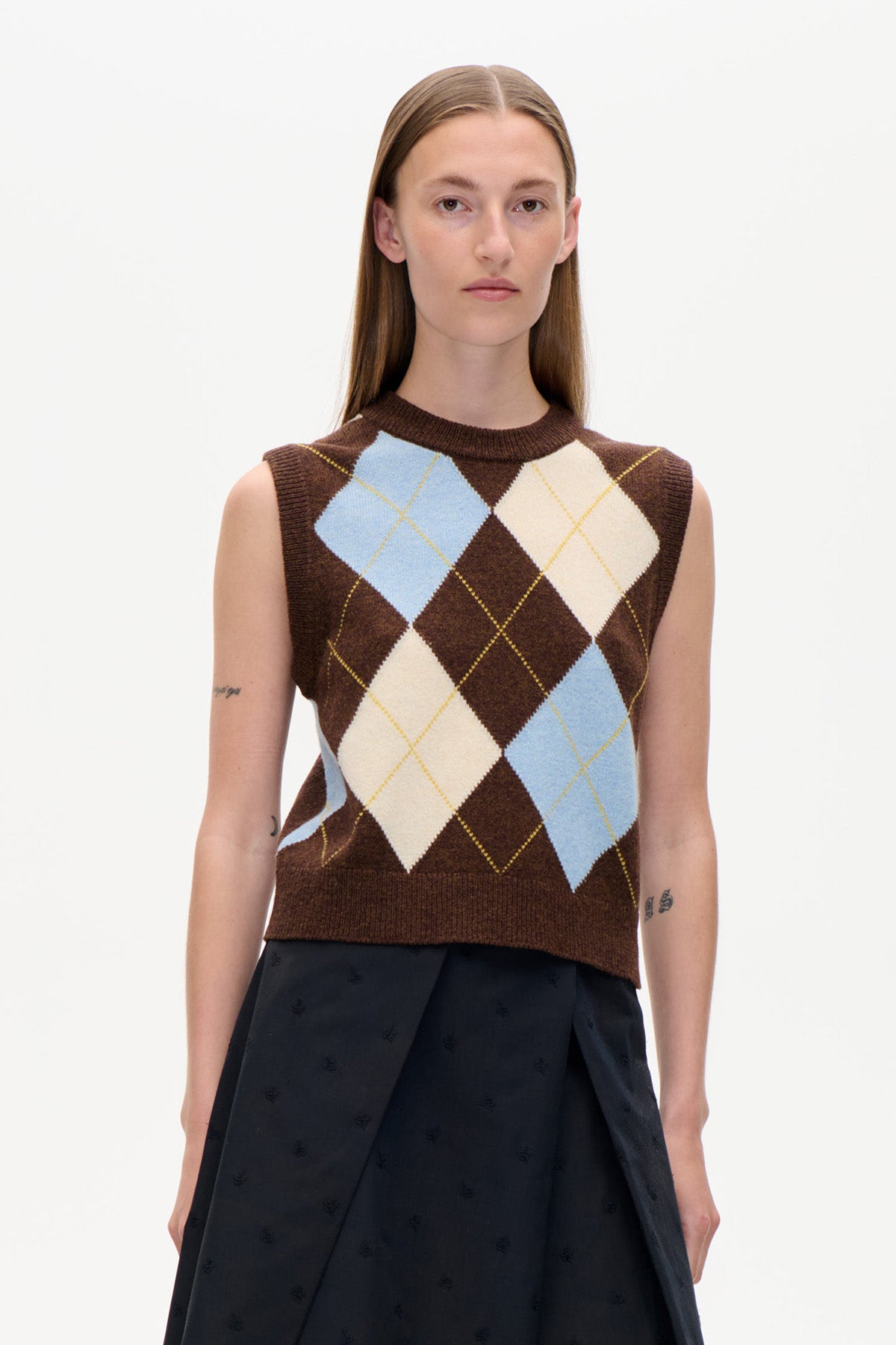 Vorderansicht einer ärmellosen braunen Strickweste mit großen hellblauen und cremefarbenen Argyle-Rauten, getragen zu einem schwarzen Rock.