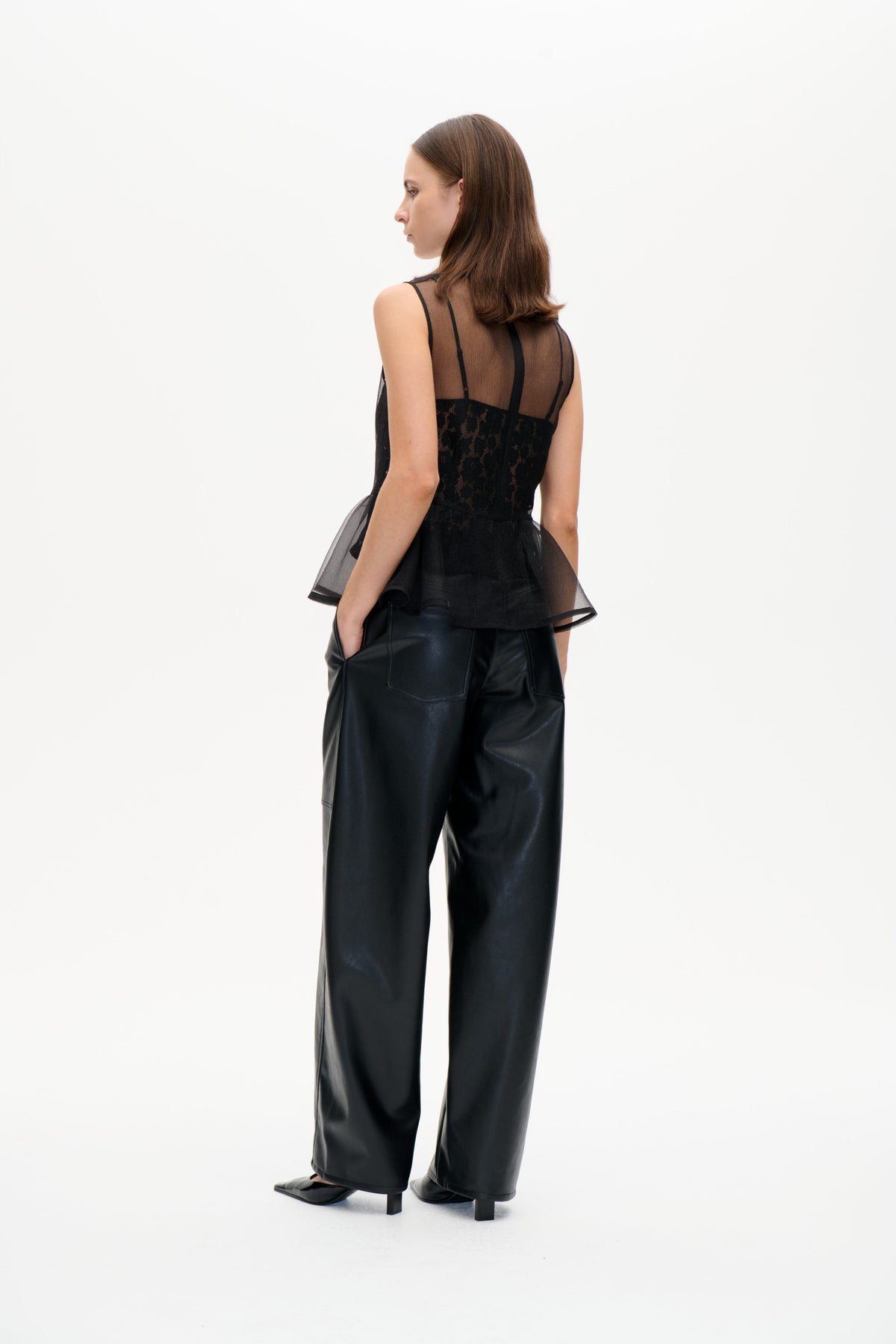 Model in einem schwarzen, transparenten, ärmellosen Top mit vertikaler Borte und Spitzeneinsatz, kombiniert mit einer weiten schwarzen Hose, von hinten.