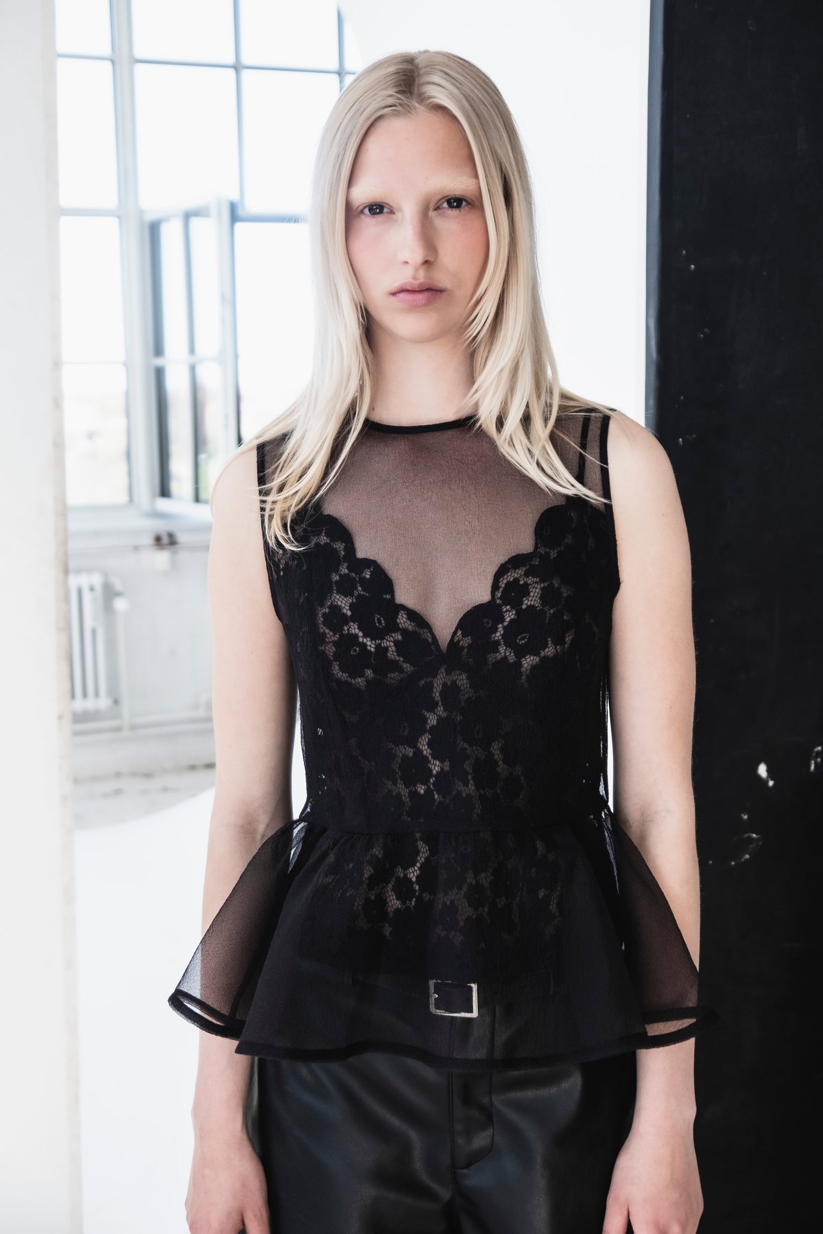 Nahaufnahme eines Kampagnen-Porträts: Ein Model trägt ein transparentes taupe-schwarzes Top mit floralen Spitzeinsätzen und kurzen Ärmeln sowie einen schwarzen Rock mit Gürtel.