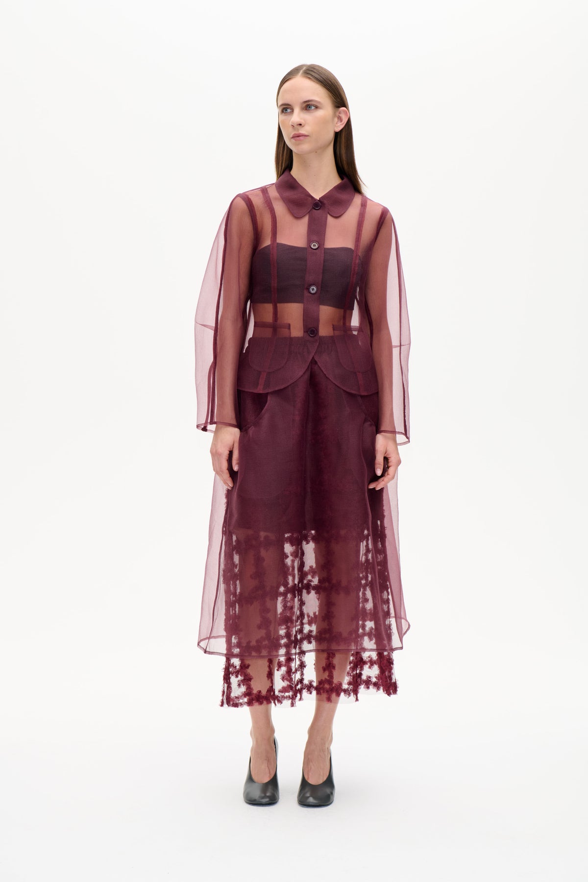 Model trägt eine transparente, pflaumenfarbene Tunika mit Knopfleiste, rundem Kragen und vertikalen Einsätzen über einem dunklen Top und einem geblümten Rock.