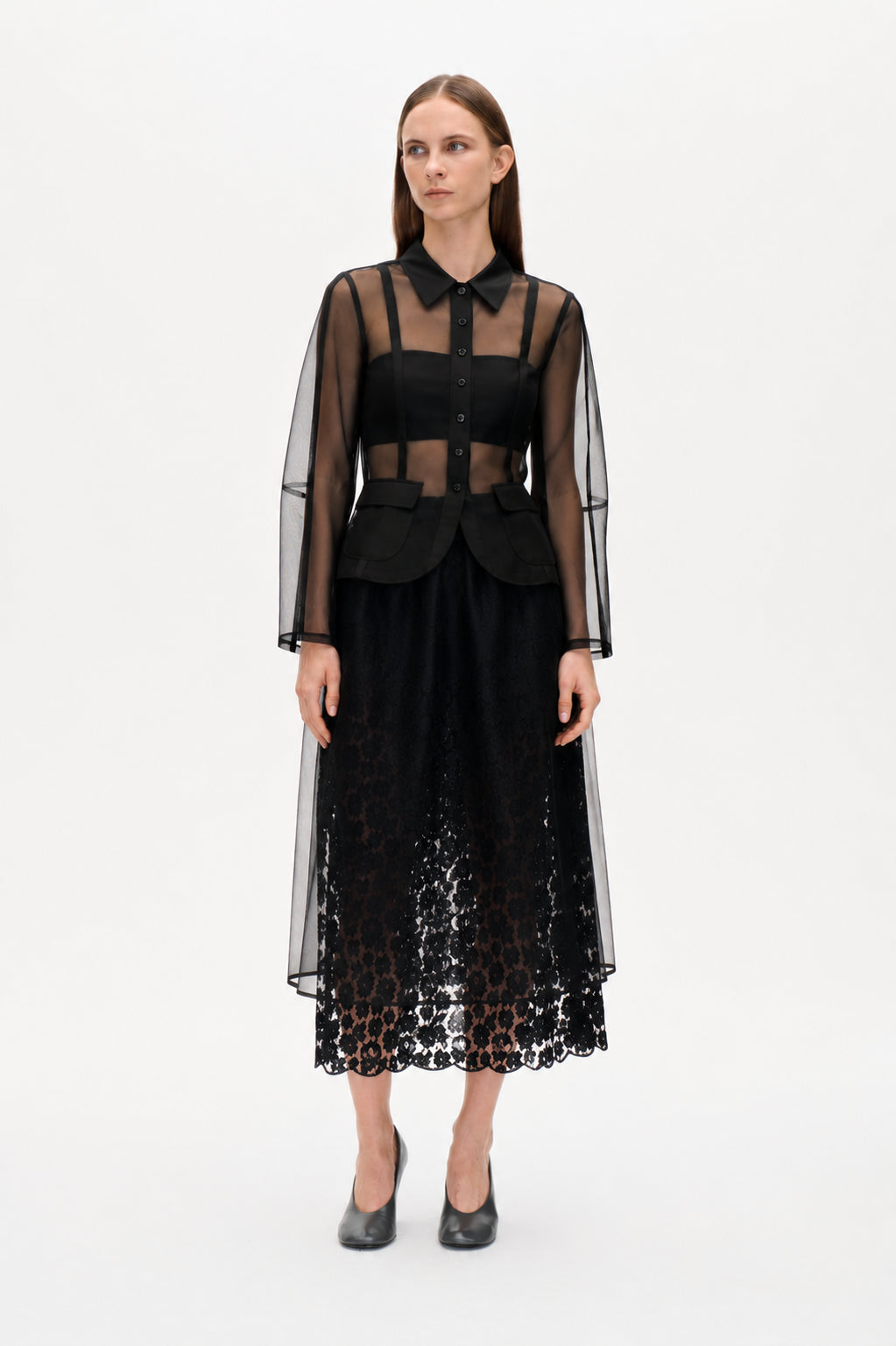 Vorderansicht einer transparenten schwarzen Bluse mit durchgehender Knopfleiste, spitzem Kragen und Pattentaschen, über einem schwarzen Top getragen, kombiniert mit einem langen Spitzenrock mit gewelltem Saum.