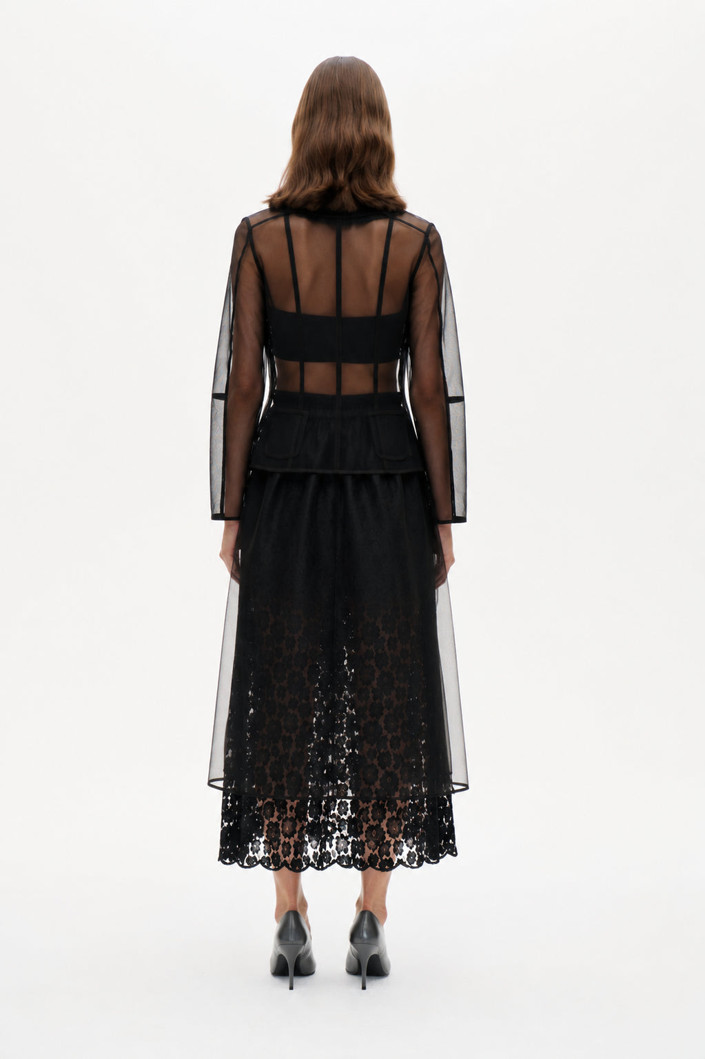 Rückansicht einer transparenten, langärmeligen schwarzen Jacke mit Teilungsnähten und Klappentaschen, die über einem schwarzen Spitzen-Midikleid getragen wird.