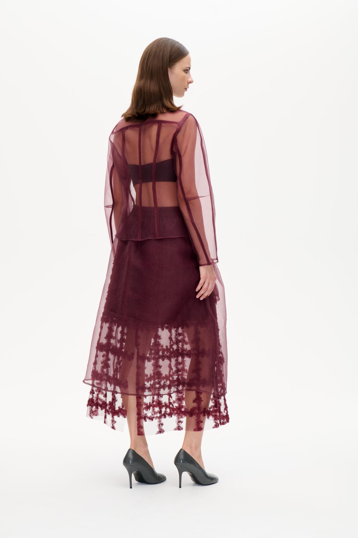 Model trägt eine transparente, pflaumenfarbene Langarmbluse mit dunklen Teilungsnähten, die über einem dunklen Outfit liegt, dazu ein transparenter Rock mit burgunderroten Blumendetails.