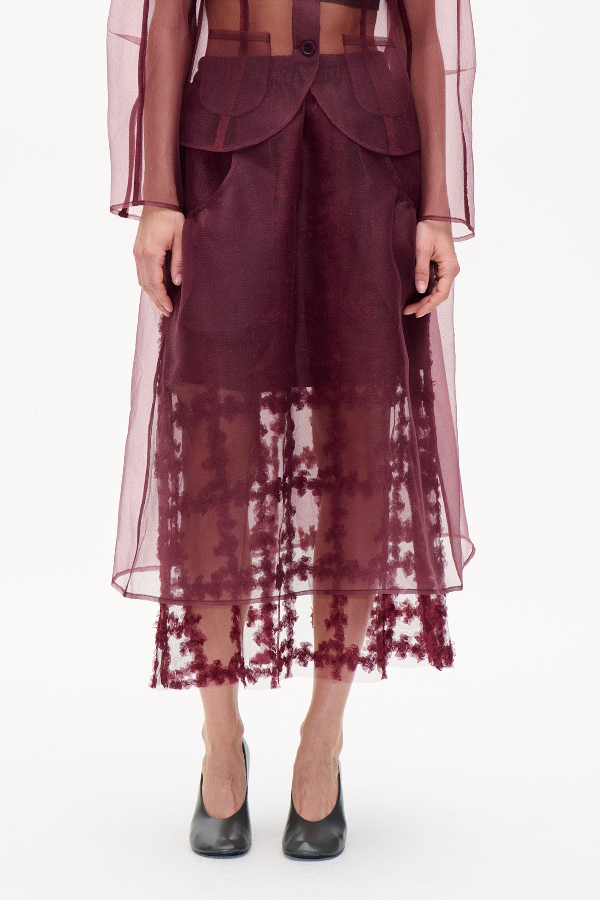 Gekürzte Vorderansicht eines Models, das einen tief bordeauxfarbenen Rock mit transparentem, geblümtem Saum und eine passende, durchscheinende Jacke mit abgerundeten aufgesetzten Taschen trägt.